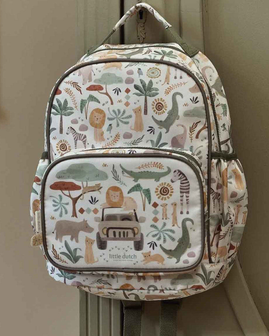 A small backpack with a safari-themed animal and vehicle print, hanging on a beige wall next to a framed picture.